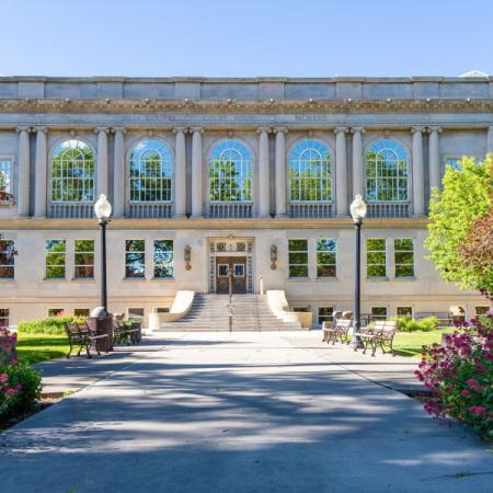 Photograph of the front of old Mesa County Courthouse in spring showing the wide staircase, four park benches, trees, flowering plants, shrubs, and grass area