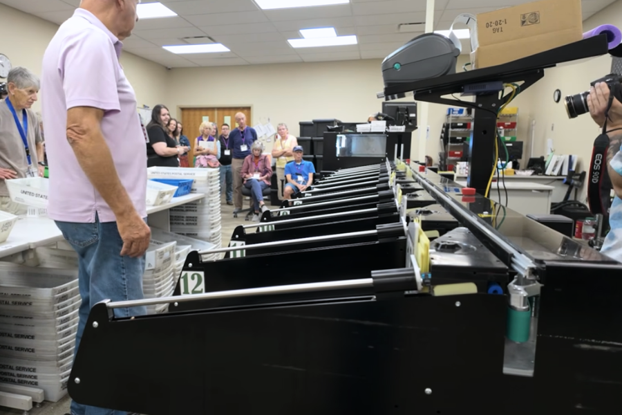 Photo of a large, black ballot sorting machine in the county elections office. About a dozen people stand around the equipment during a tour of the Mesa County elections department.